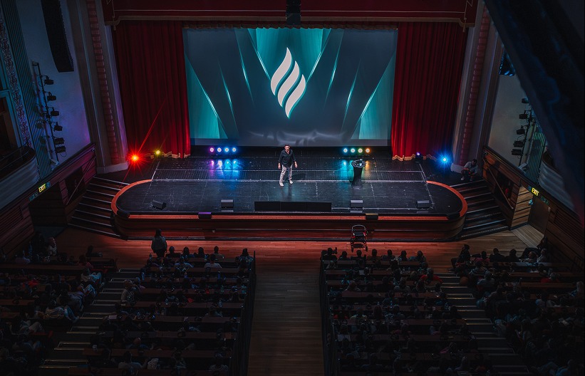 Alexis Alcalá on stage at a packed theater with the InnerCamp logo projected behind him