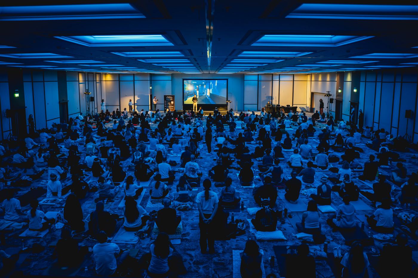 Wide shot of a packed ballroom audience seated on mats in deep blue ambient light at an InnerCamp event